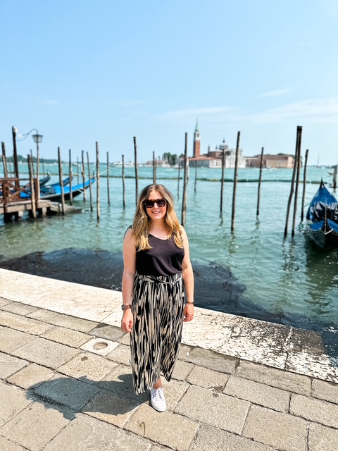 Laura stands on the edge of a path in Venice overlooking the boats that line the walkway. It's a beautiful warm day with clear blue skies.